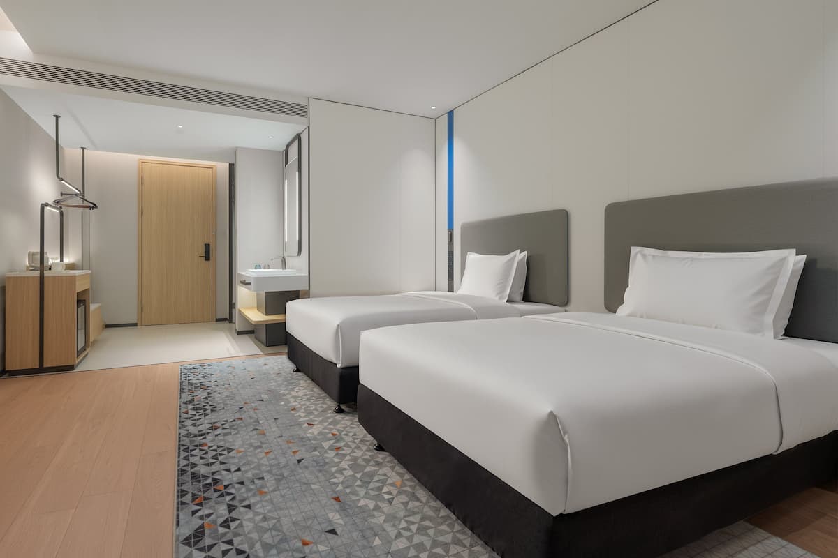 A modern hotel room with two beds, a large mirror, a wooden wardrobe, and a bathroom with a sink and mirror.