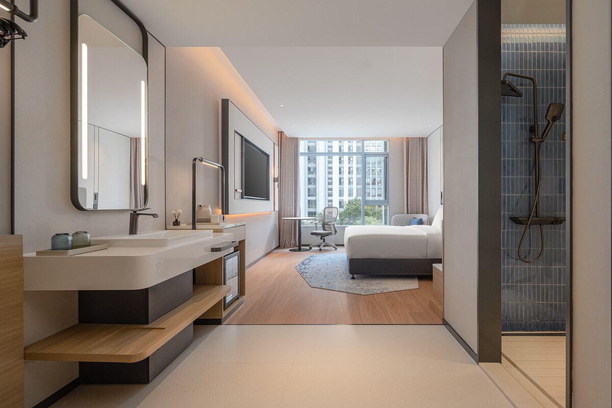 A modern hotel bathroom with a large mirror, dual sinks, a shower, and a bed with a view of the city.