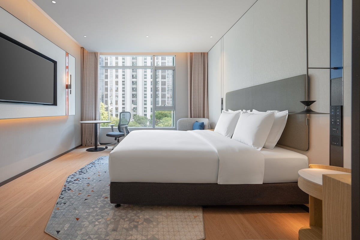 A modern hotel room with a large bed, a flat-screen TV mounted on the wall, a small table, and a chair.