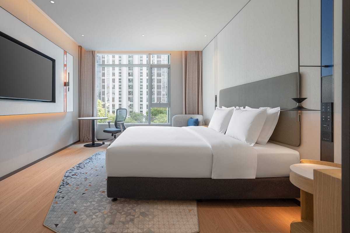 A modern hotel room with a large bed, a flat-screen TV mounted on the wall, a small table, and a chair.