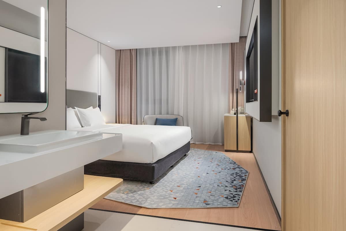 A modern hotel room with a large bed, a bedside table, a built-in TV, and a large mirror.