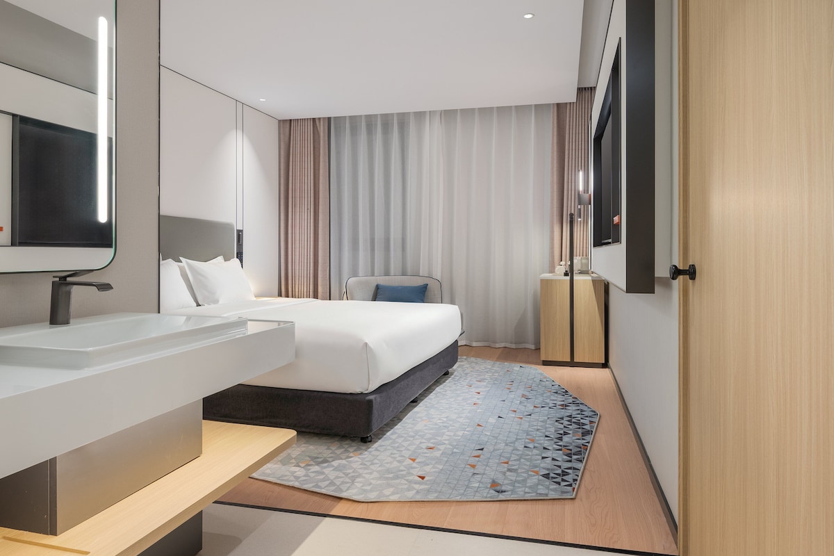 A modern hotel room with a large bed, a bedside table, a built-in TV, and a large mirror.