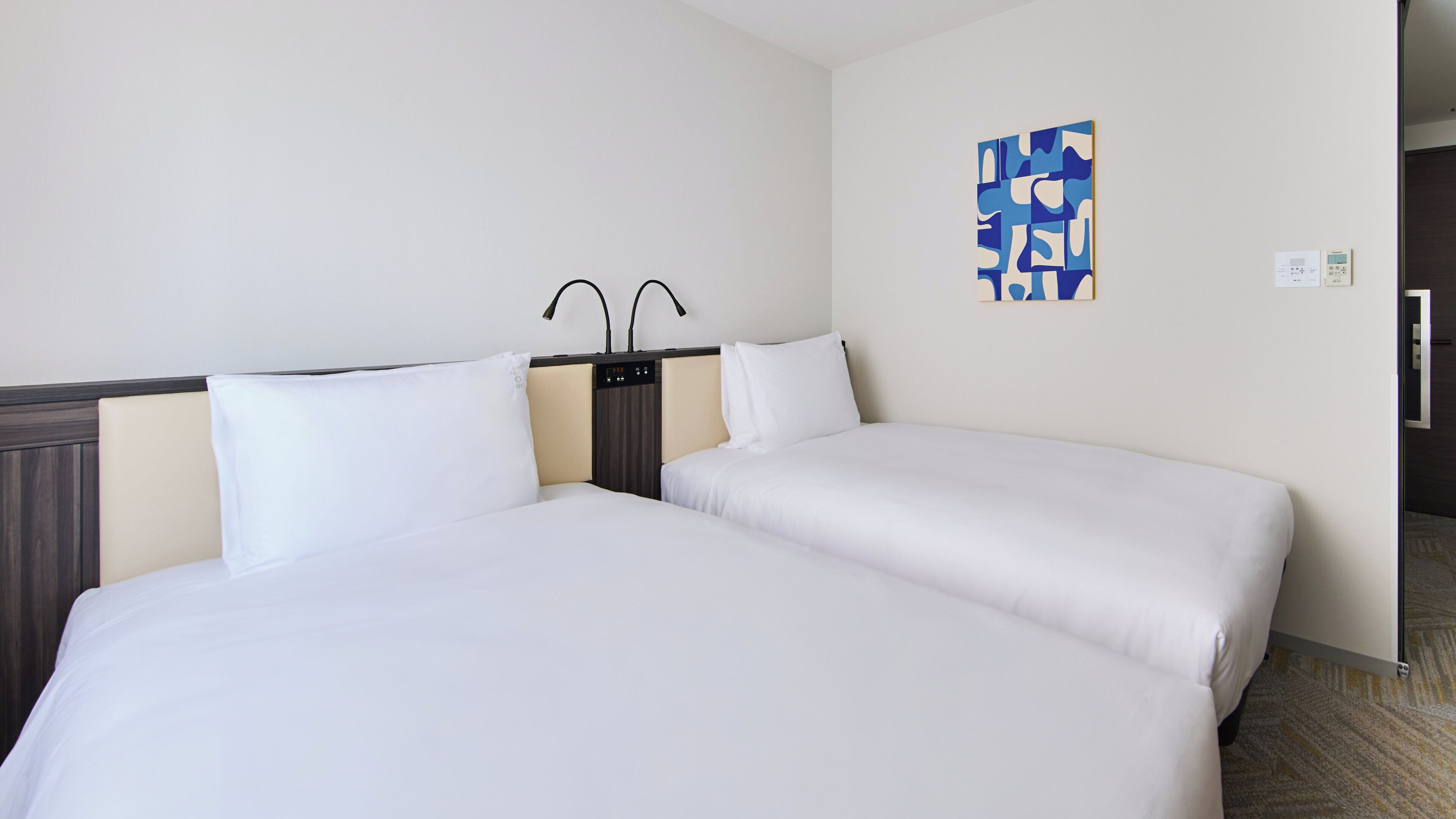 Une chambre d’hôtel avec deux lits, une table de chevet avec une lampe, une œuvre d’art accrochée au mur et une porte.