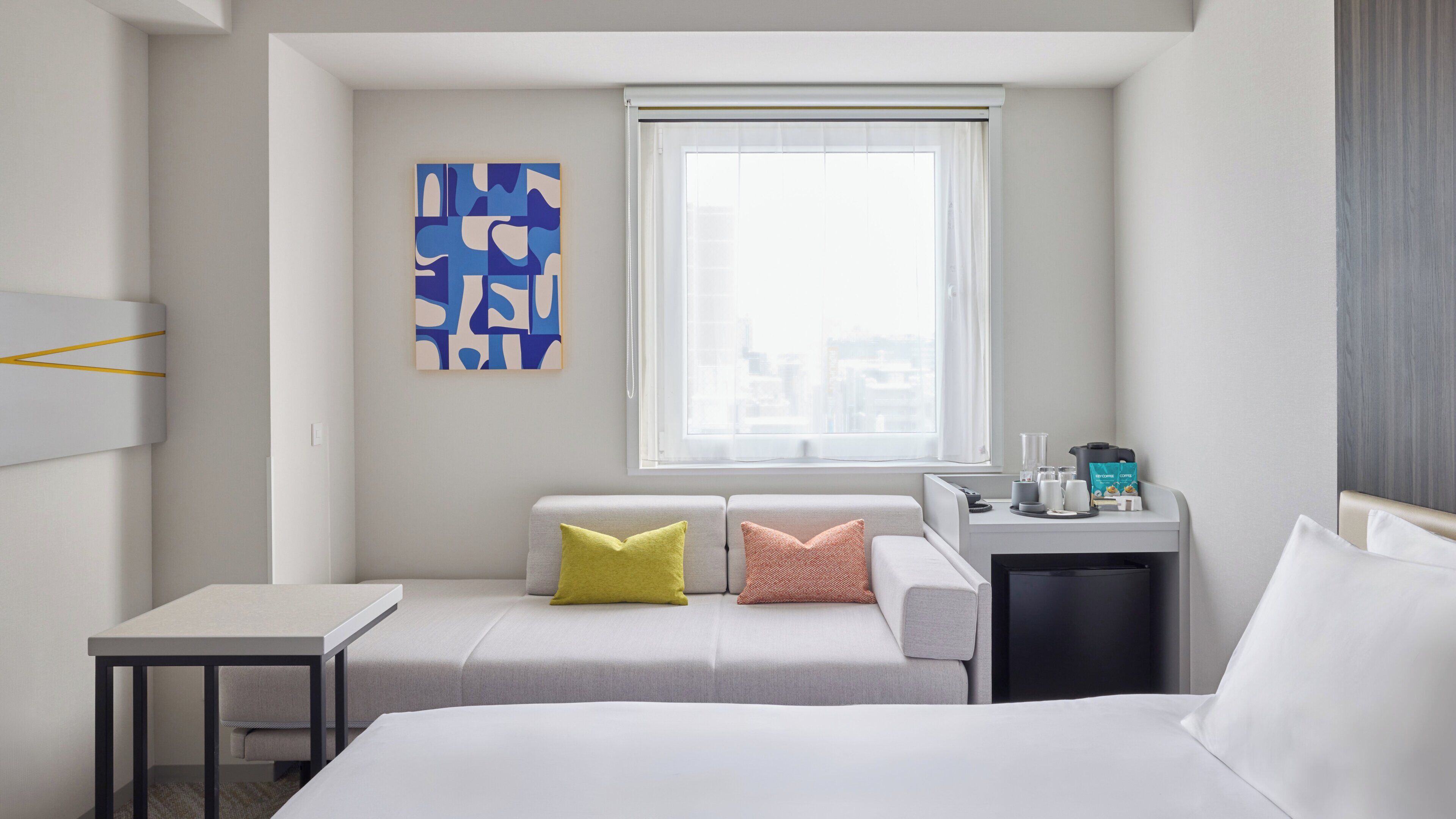 Hotelkamer met een bed, een klein bureau, een raam met uitzicht, een schilderij aan de muur en een nachtkastje met een koffiezetapparaat.