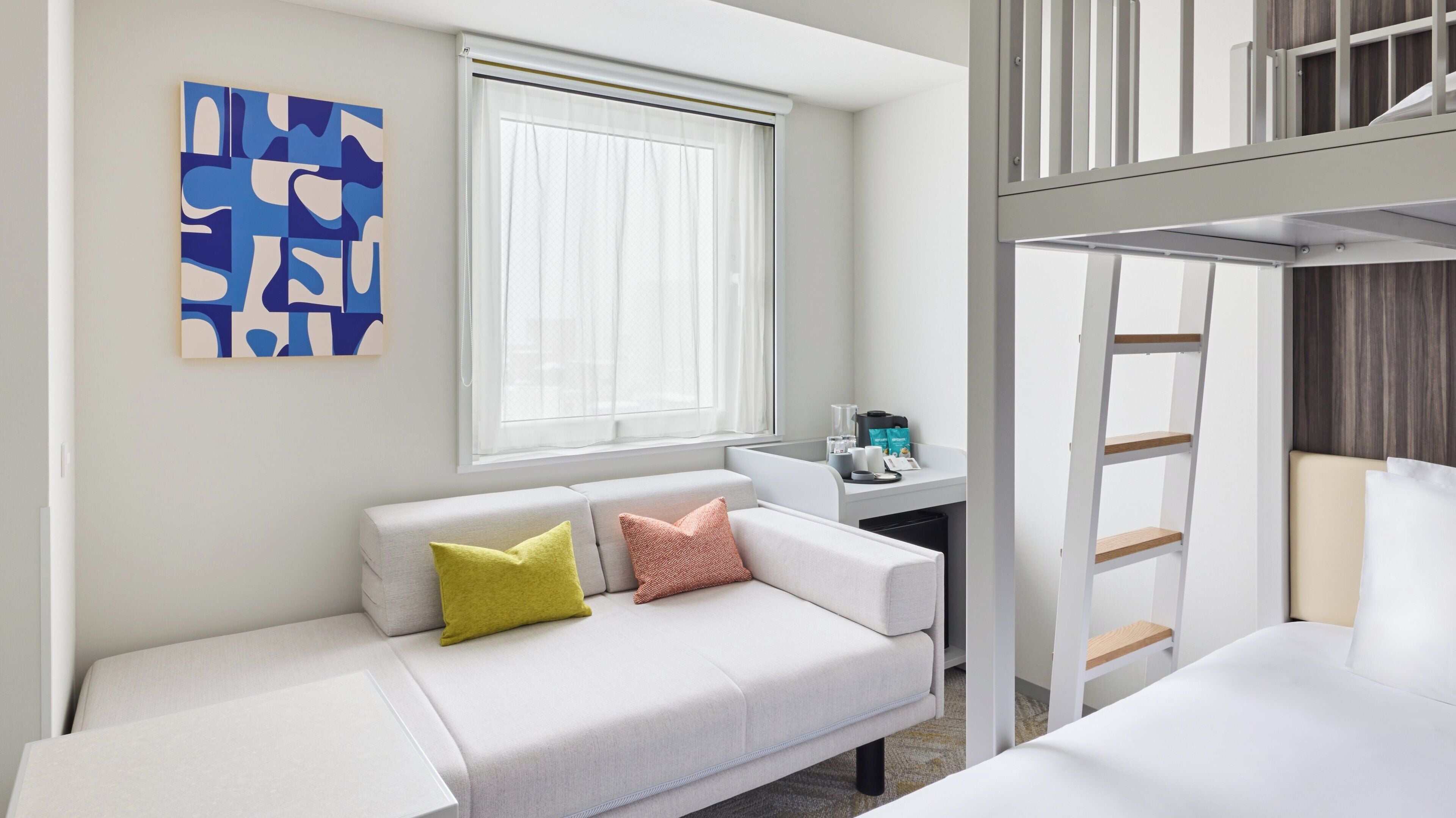 Ein Zimmer mit einem weißen Sofa, einem Schreibtisch mit Computer, einem Fenster mit durchsichtigen Vorhängen und einem Etagenbett.
