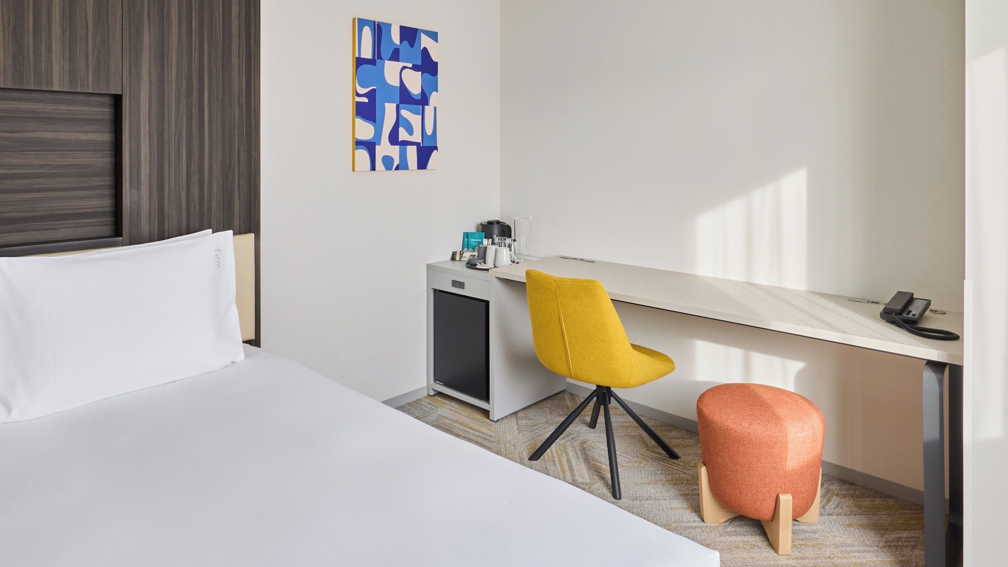 Una habitación de hotel con cama, escritorio, silla, un mueble bajo y una obra de arte en la pared.
