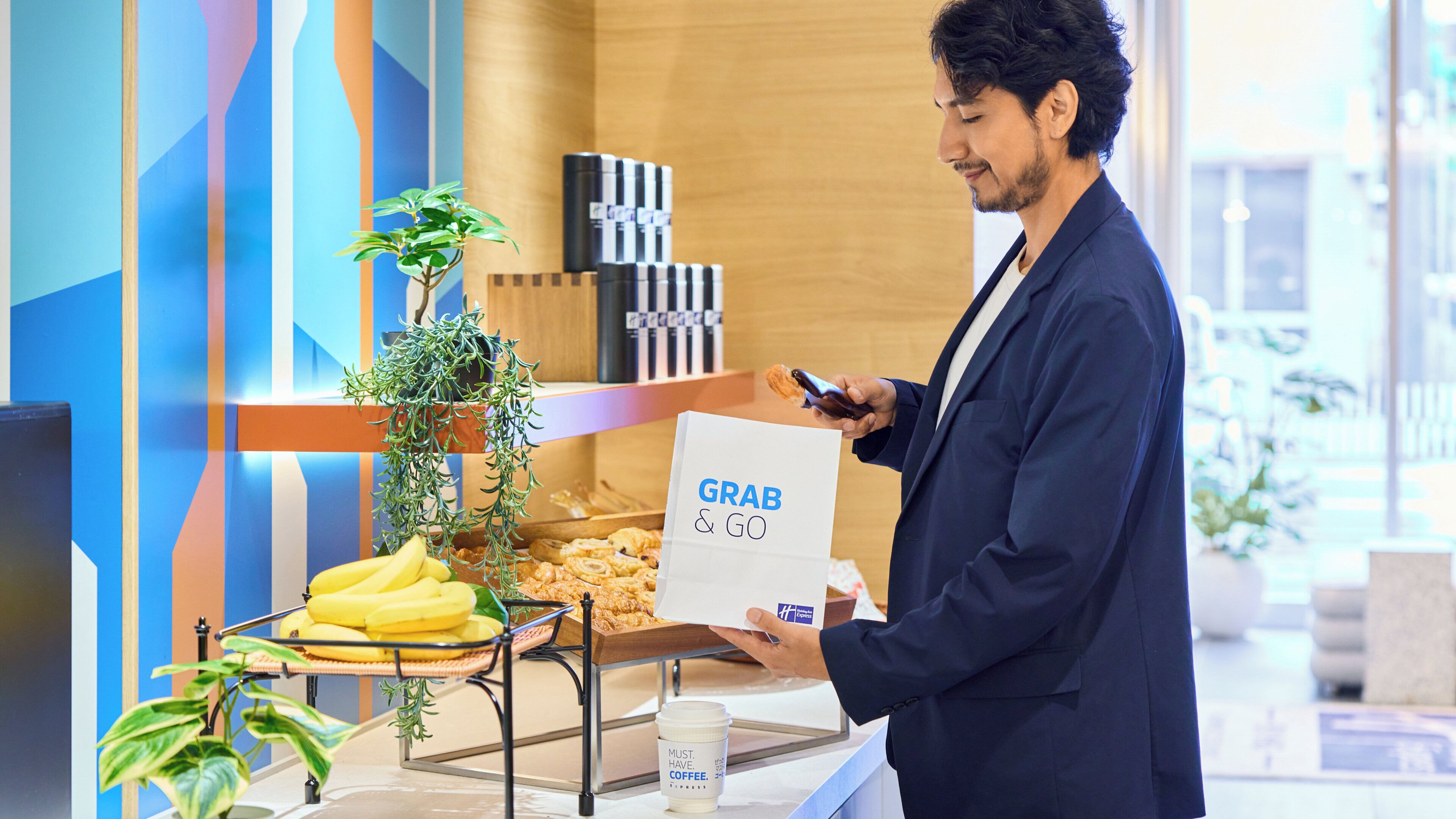 A person holding a 'GRAB & GO' bag at a counter with a coffee cup and a display of food items.