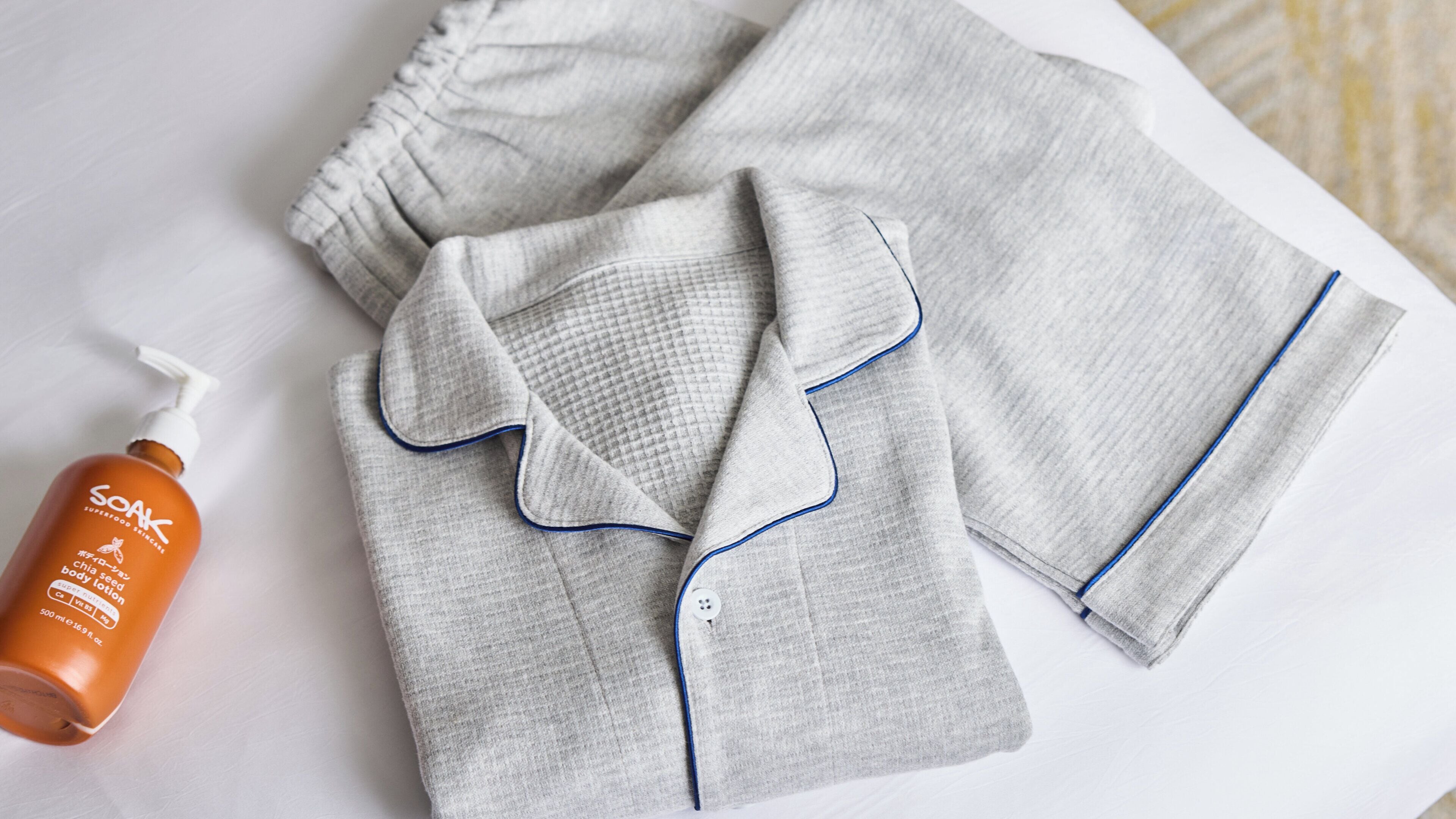 Ein ordentlich gefaltetes, graues Pyjama-Set mit blauen Akzenten, eine Flasche Soak Körperlotion und eine weiße Fläche.