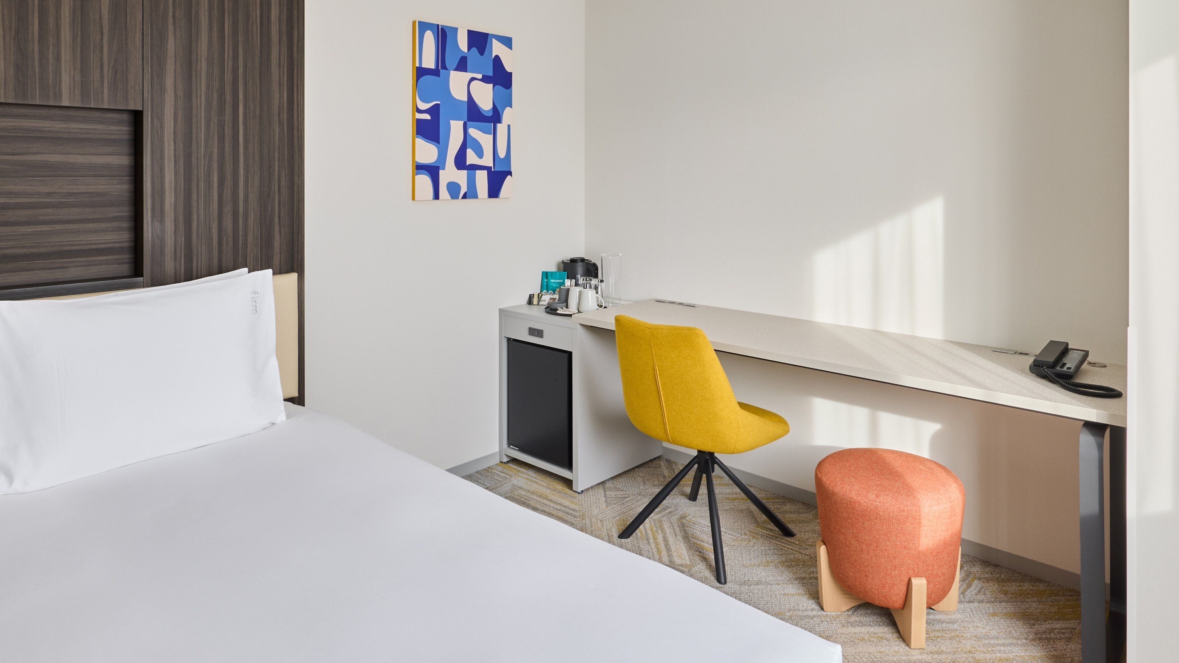 A hotel room with a bed, a desk, a chair, a small refrigerator, a coffee maker, and a phone.