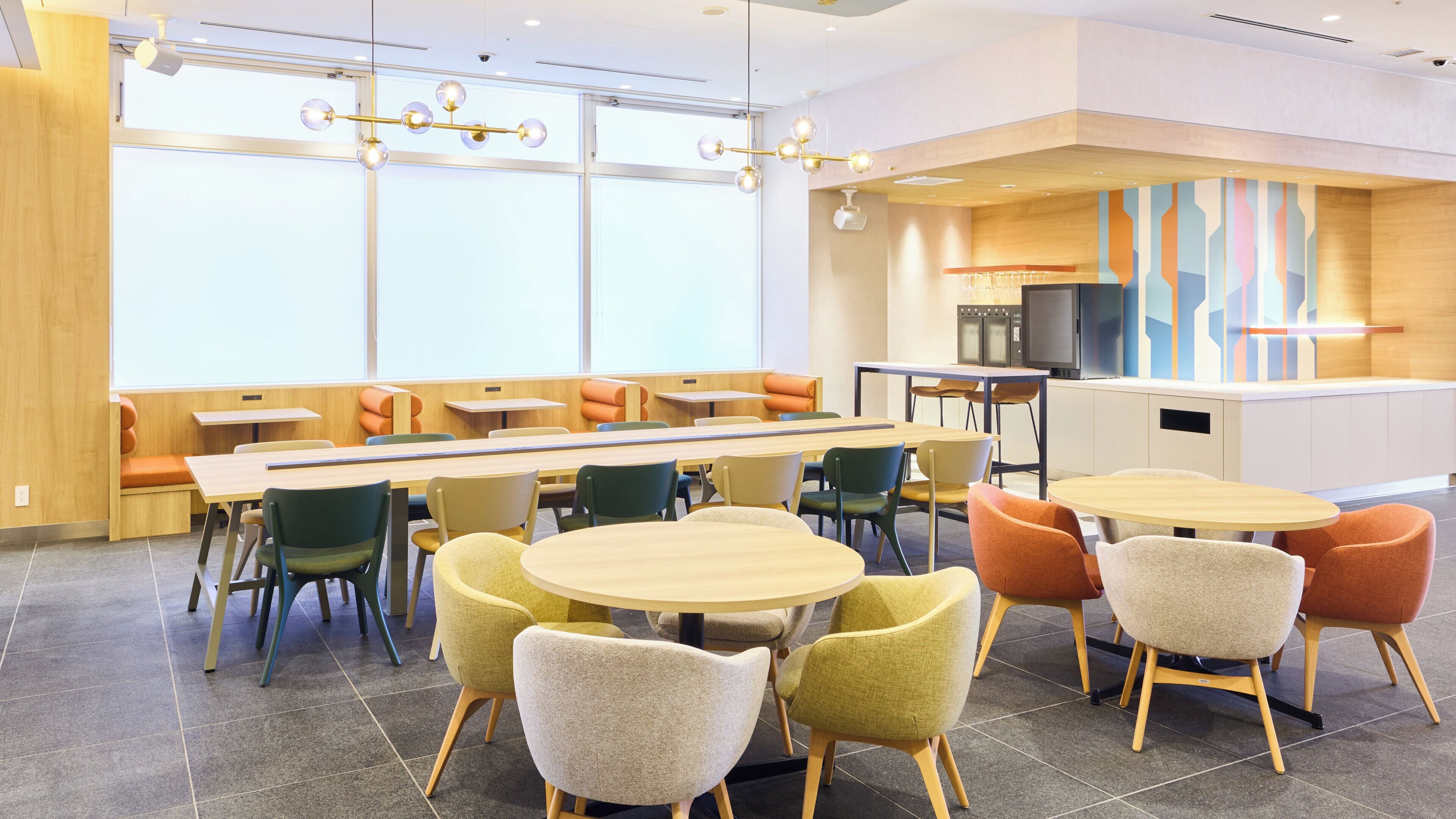 A modern dining area with long tables and chairs, a kitchen area with a microwave, and a large window providing natural light.