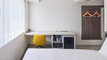 A hotel room with a bed, a desk with a chair, a wardrobe, and a window with blinds.