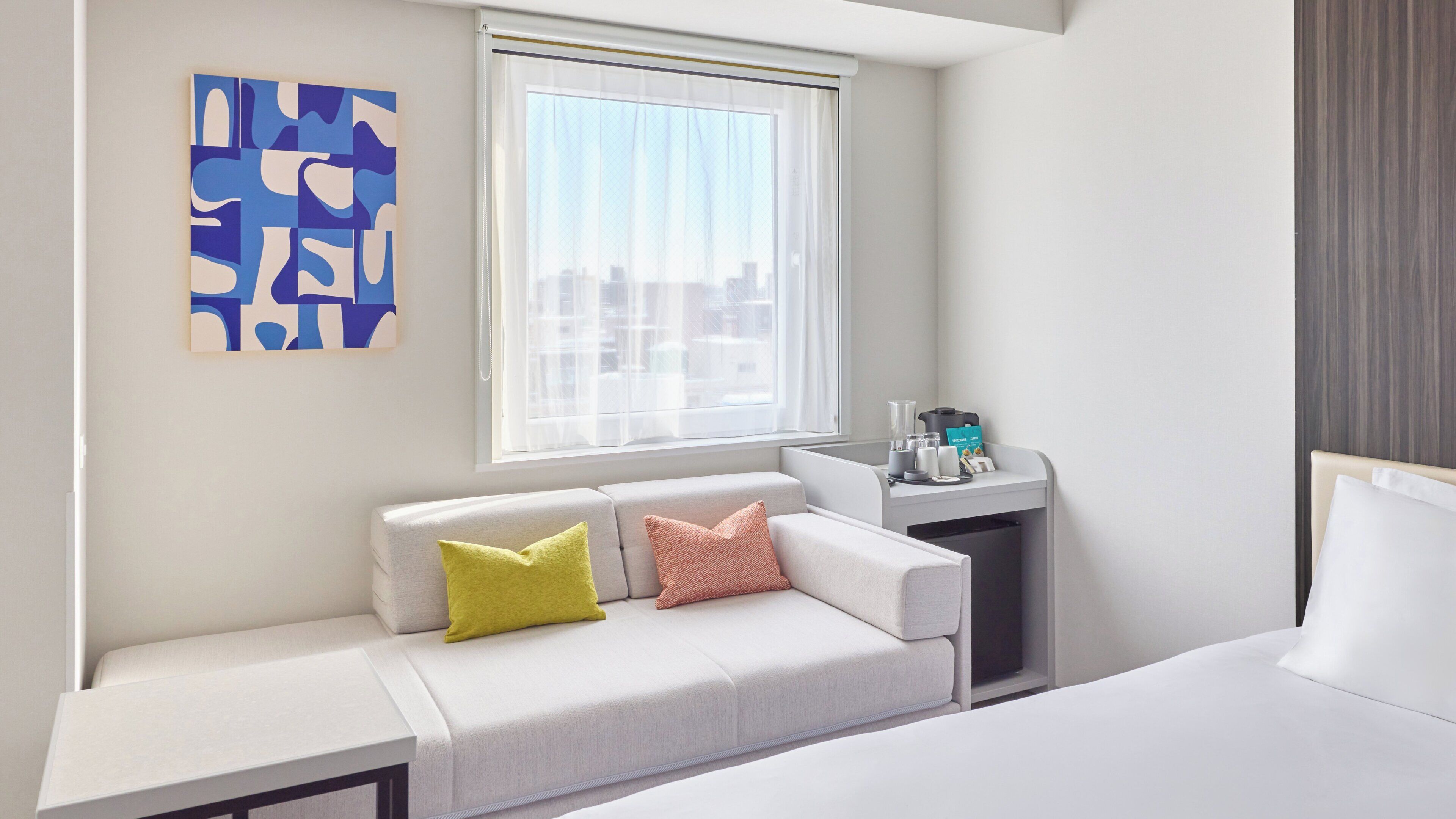 A hotel room with a bed, a sofa, a small table with a coffee maker, and a window with a view of buildings.
