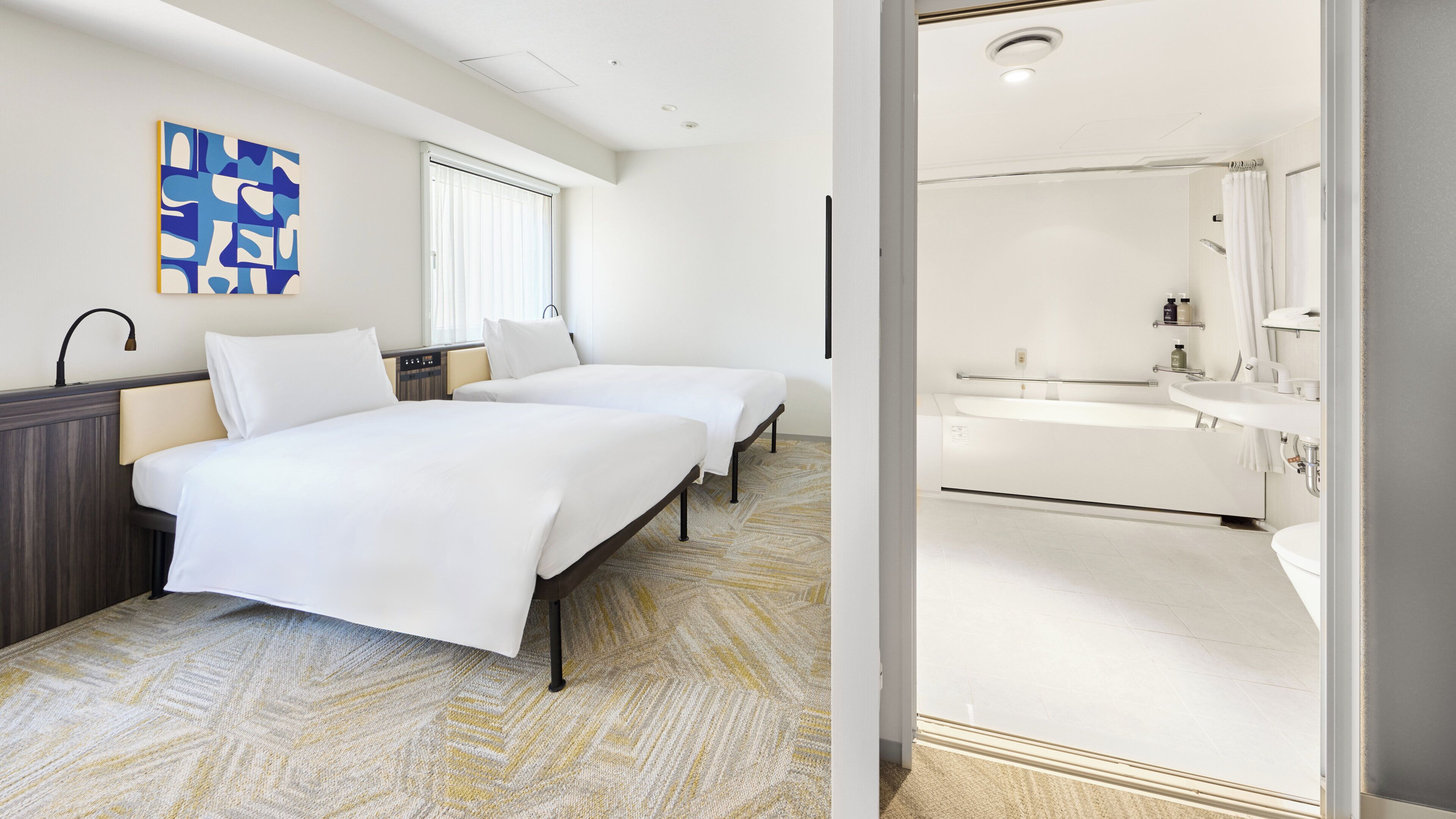 Een hotelkamer met twee bedden, een groot raam, een badkamer met een wastafel en spiegel, en een blauw abstract schilderij aan de muur.