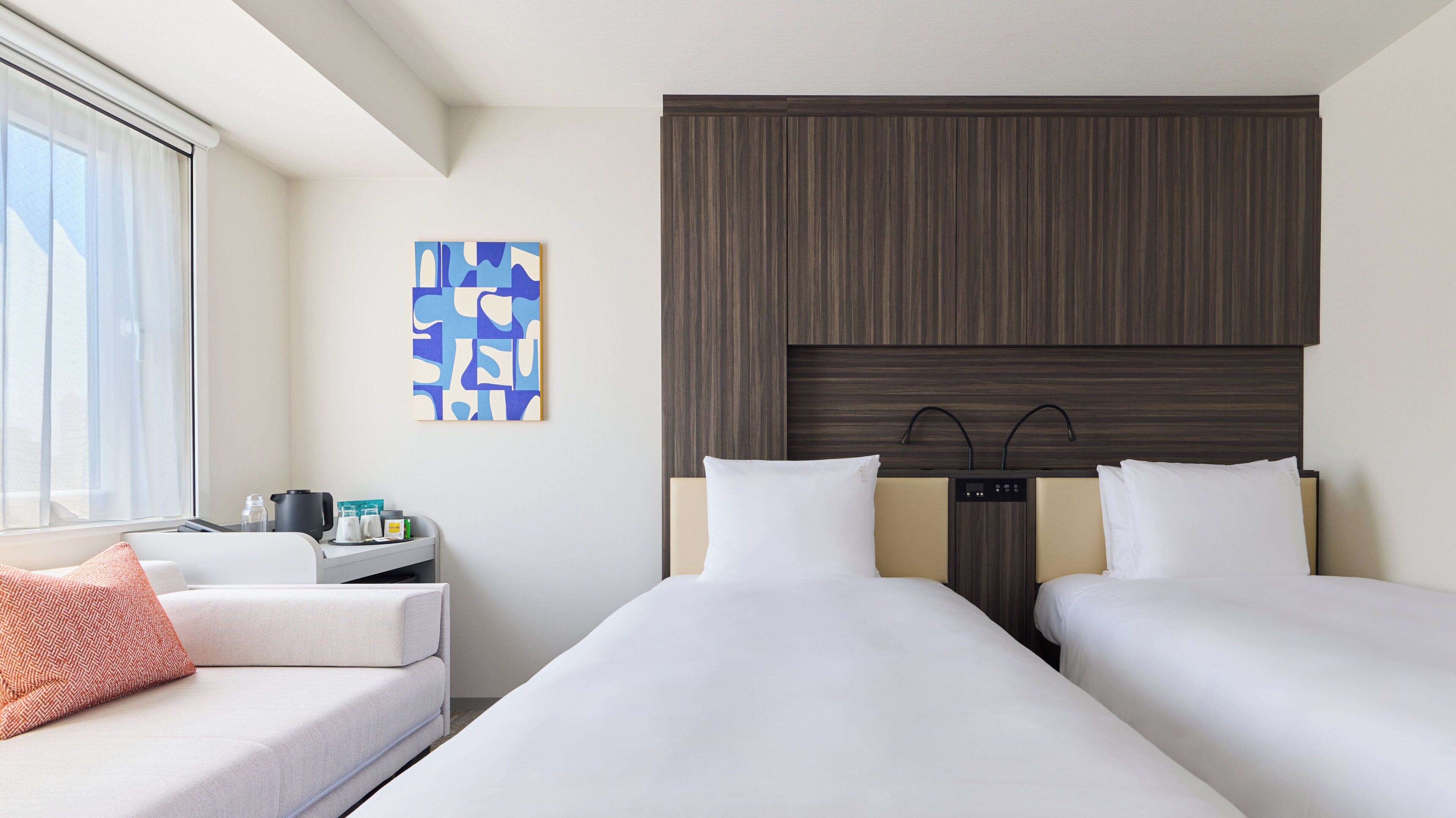 A hotel room with two beds, a bedside table with a lamp, a coffee maker, a small table with a bottle and a cup, and a painting on the wall.