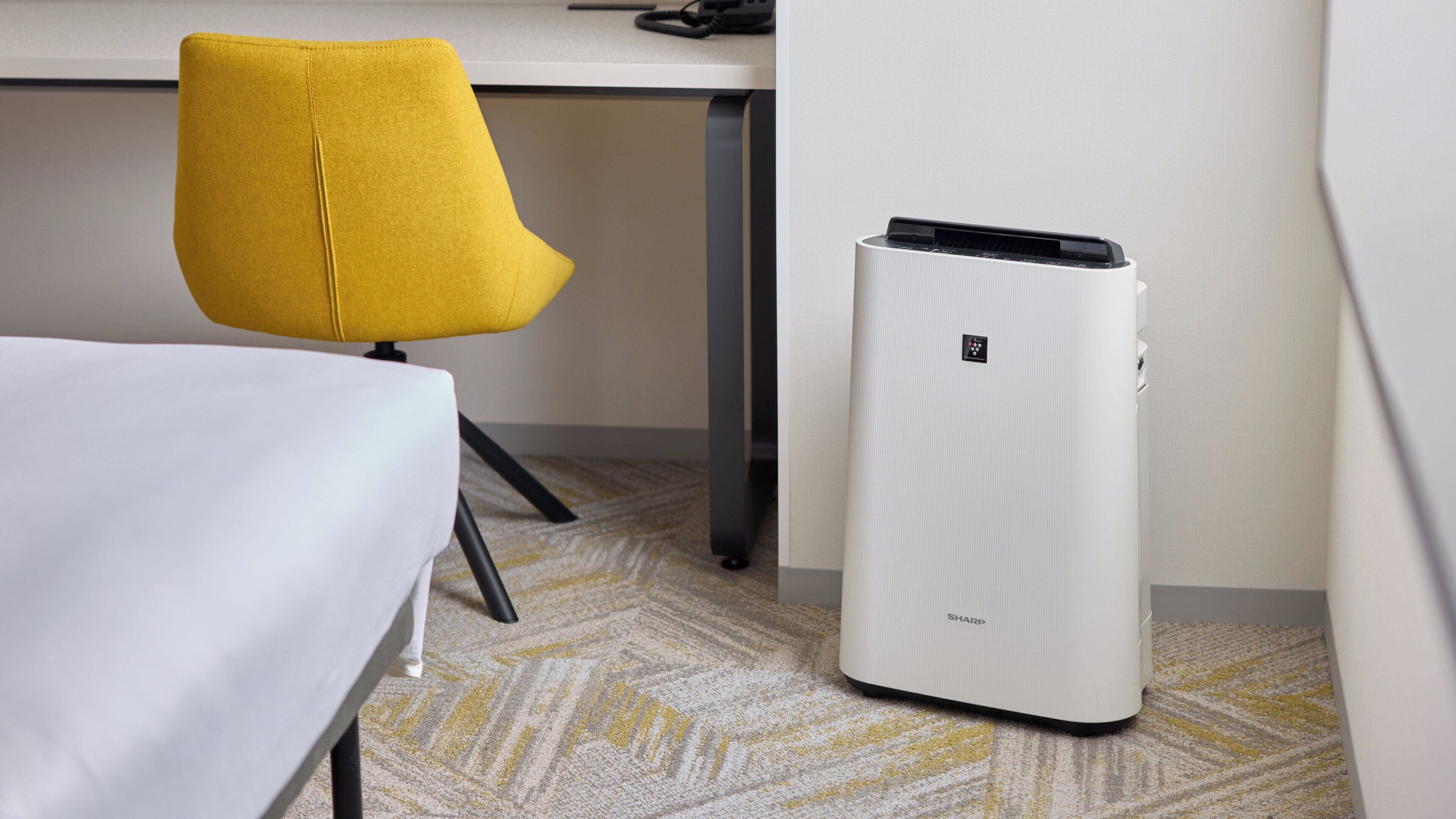 A hotel room with a bed, a yellow chair, a desk, and a white air purifier.