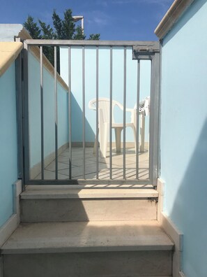 Una entrada con reja a un balcón con escalones, una silla de plástico blanca y un cielo despejado al fondo.