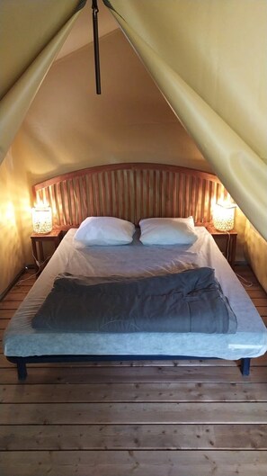 Un lit simple dans une chambre amĂ©nagĂ©e comme une tente, avec une tĂȘte de lit en bois, deux lampes de chevet et une couverture sur le lit.