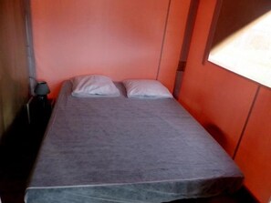 Un lit simple avec un matelas et des oreillers gris, une table de chevet avec une lampe et une fenêtre avec des stores.