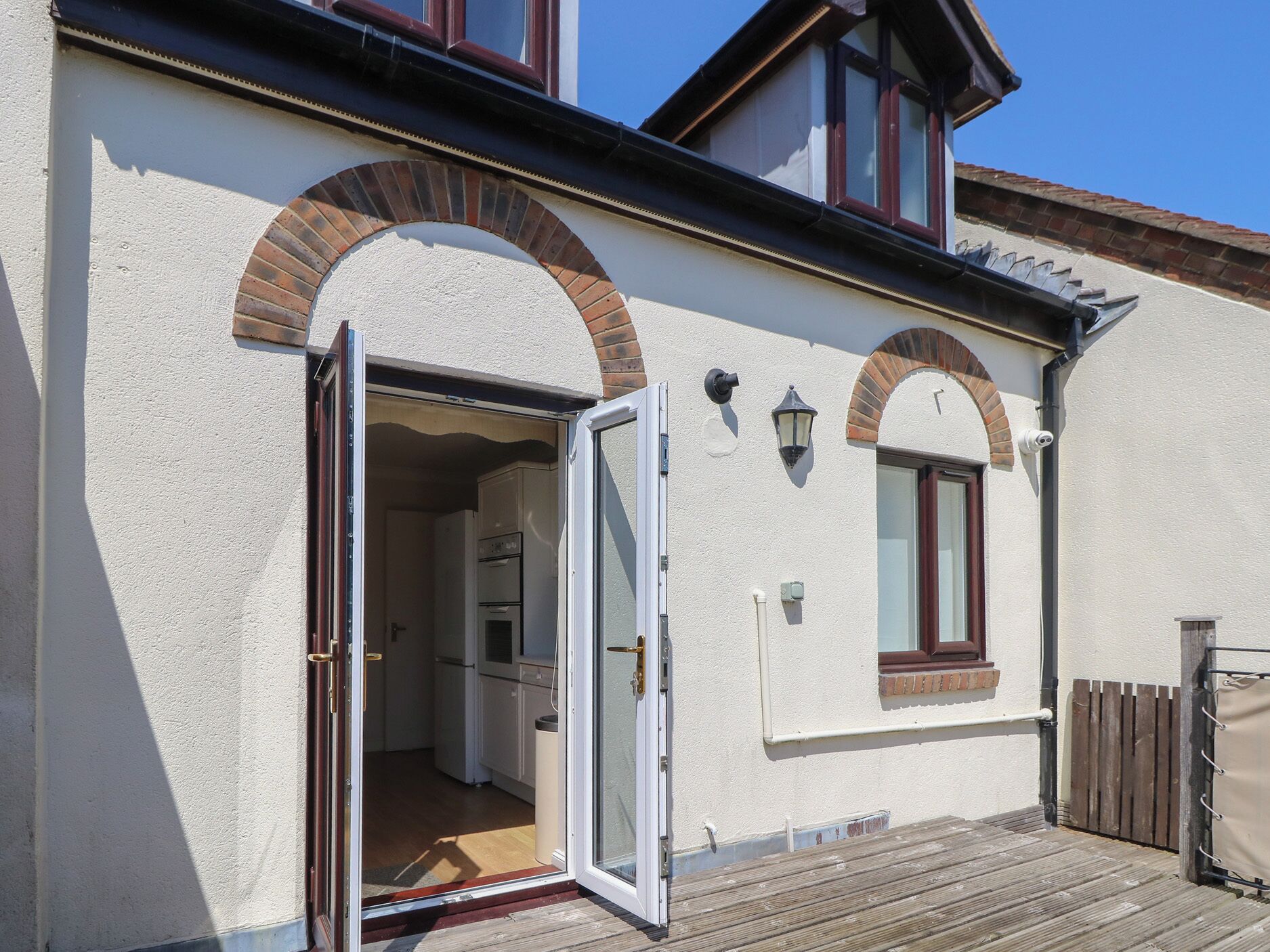 Ein Wohnhaus mit weißer Fassade, zwei Rundbogenfenstern, einer Glastür zum Verschieben, einer kleinen Küche und einer Holzterrasse.