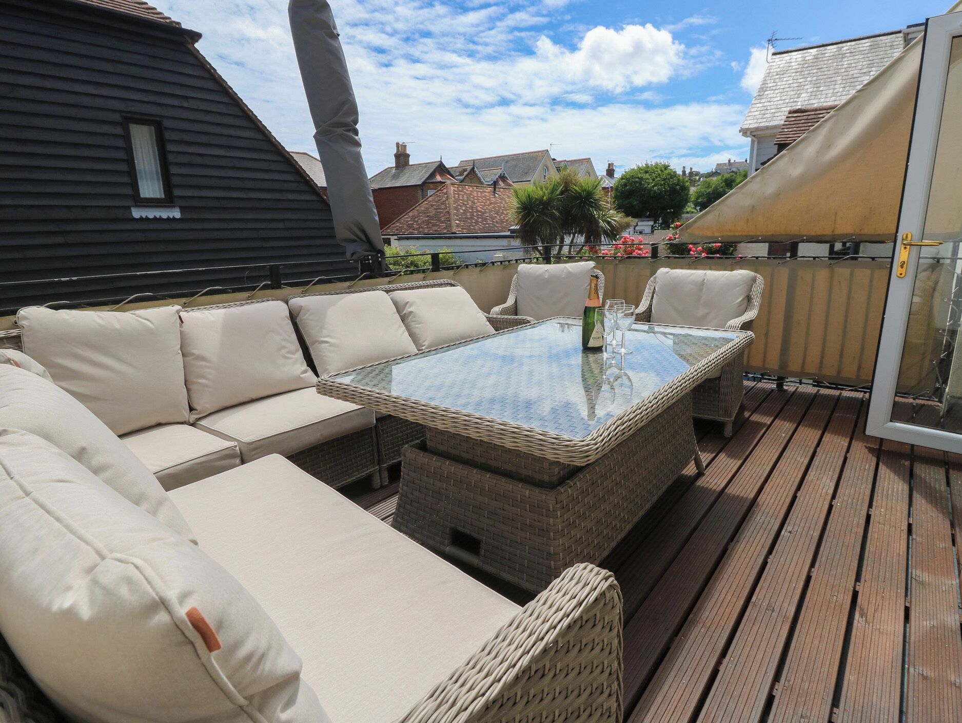 Een terras buiten met rieten meubels, een glazen tafel en een fles champagne.