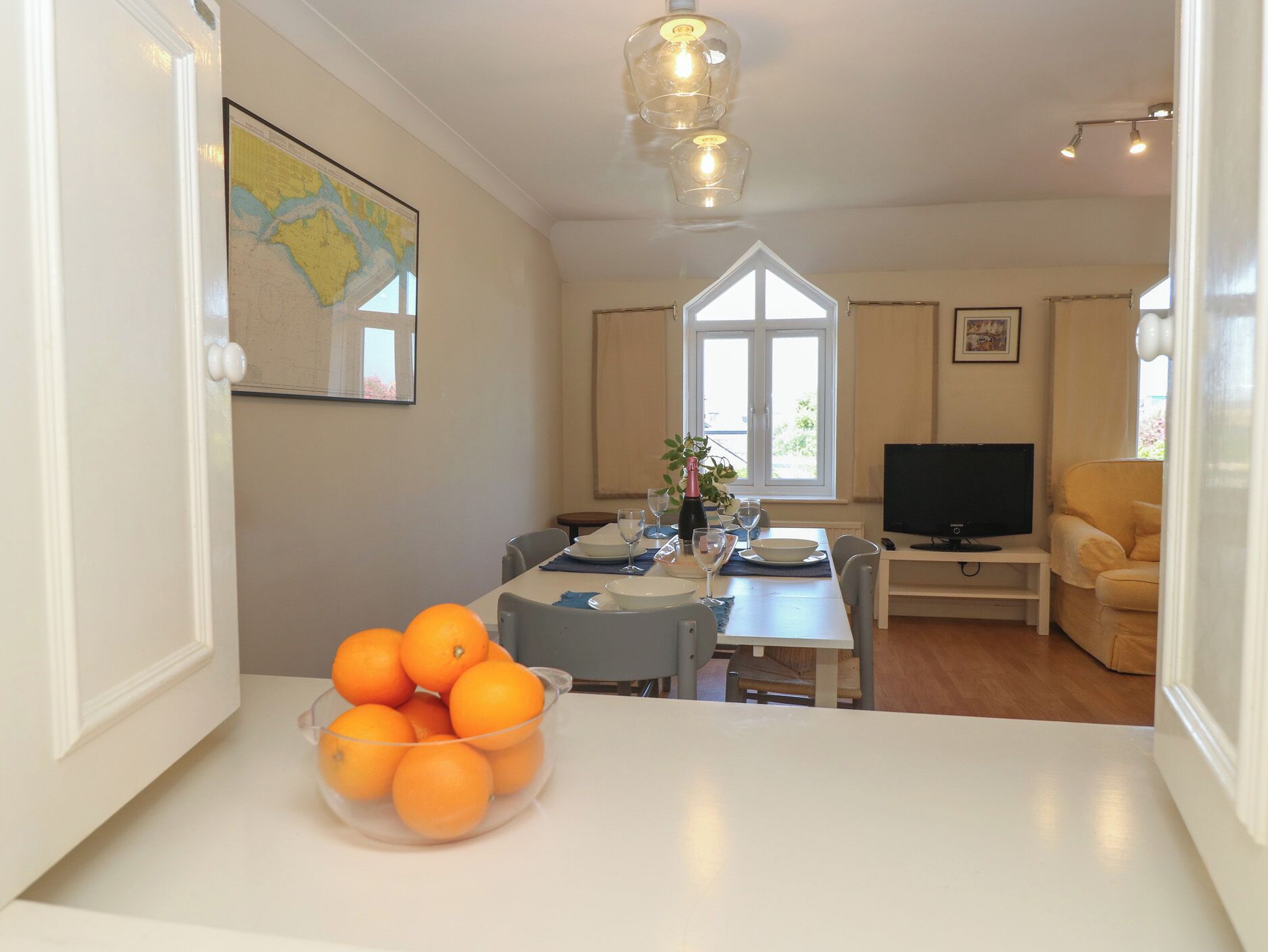 A dining area with a table set for two, a bowl of oranges, a bottle of champagne, and a television on a stand.
