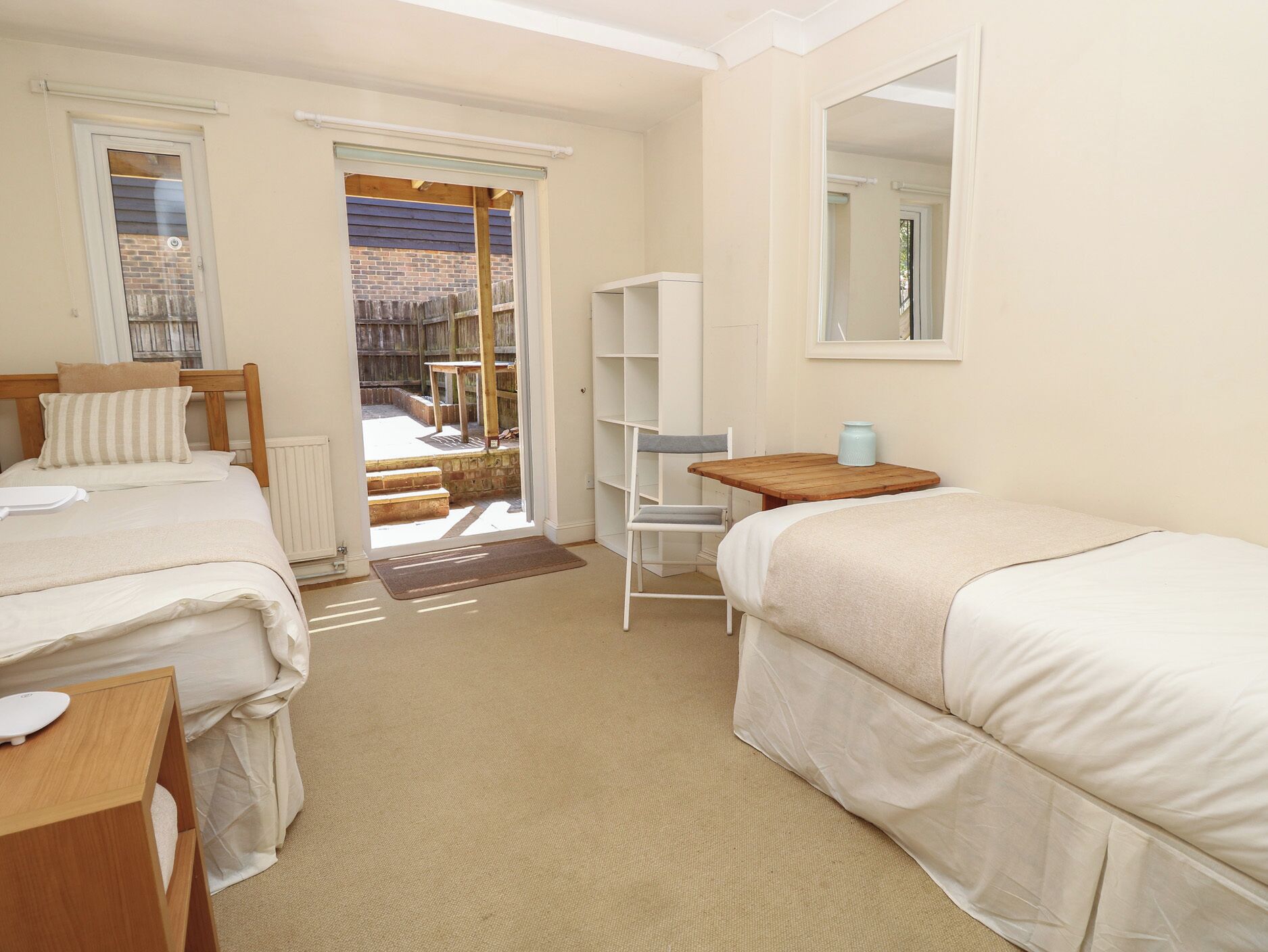 Une chambre avec deux lits, une table en bois, une chaise, une bibliothèque et une porte donnant sur un espace extérieur.