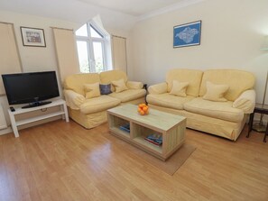 A living room with a sofa set, a television on a stand, a coffee table with books and oranges, and a window with curtains. - Anchorage House (Seaview)