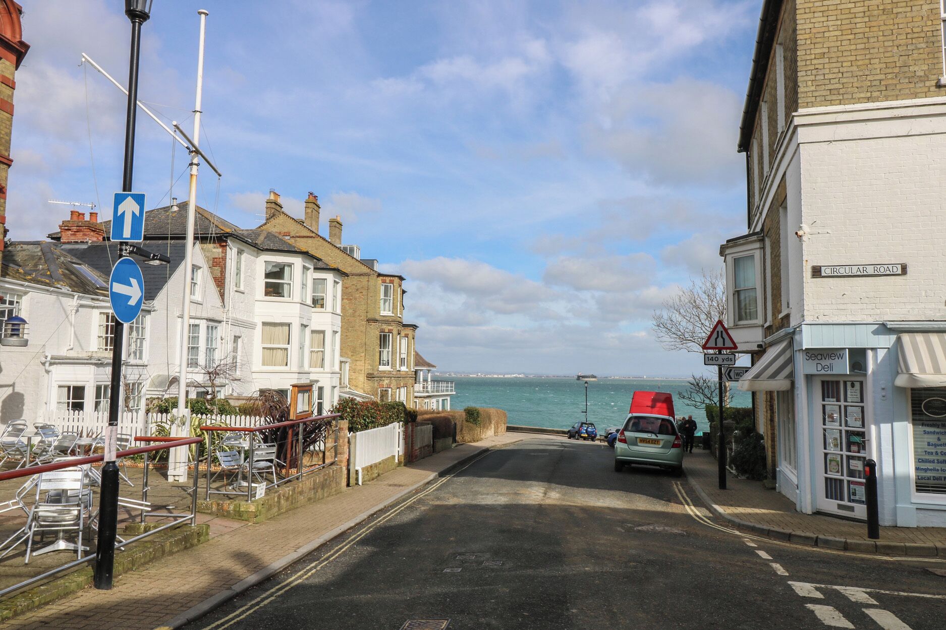 A coastal street with a view of the sea, a red car, and a Seaview Deli.