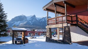 A snow-covered outdoor area with a wooden building, a stone structure, and a mountain backdrop. - Cozy Hotel Room w/ Full Kitchen | Hot Tub & Sauna (Canmore)