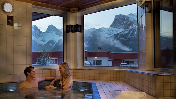 A couple in a hot tub with a mountain view. - Cozy Hotel Room w/ Full Kitchen | Hot Tub & Sauna (Canmore)