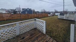 Un patio trasero con terraza de madera, cerca blanca y una casa azul al fondo.