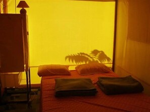 Ein Bett mit Kissen und einer zusammengefalteten Decke, ein Nachttisch mit einer Lampe und ein Fenster mit Blick auf Palmen.