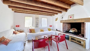Una habitación con techo de madera, chimenea, cama con almohadas, mesa con sillas y obras de arte en las paredes.