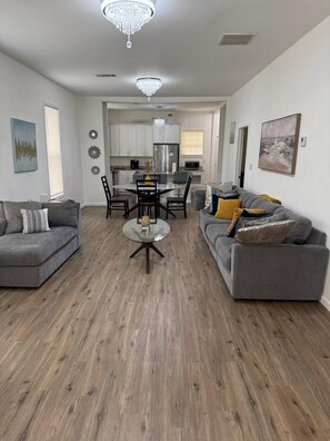 Ein modernes Wohnzimmer mit zwei grauen Sofas, einem Glastisch, einem Essbereich mit einer Pendelleuchte und einer Küche im Hintergrund.