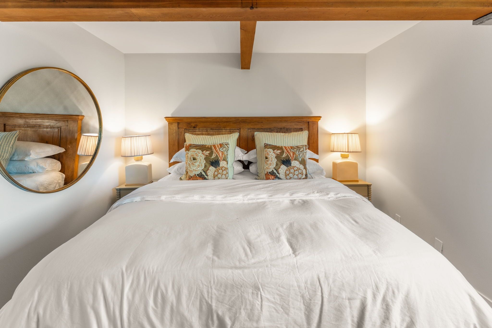 Quarto com cama grande, duas luminárias de cabeceira, espelho redondo e cabeceira de madeira.