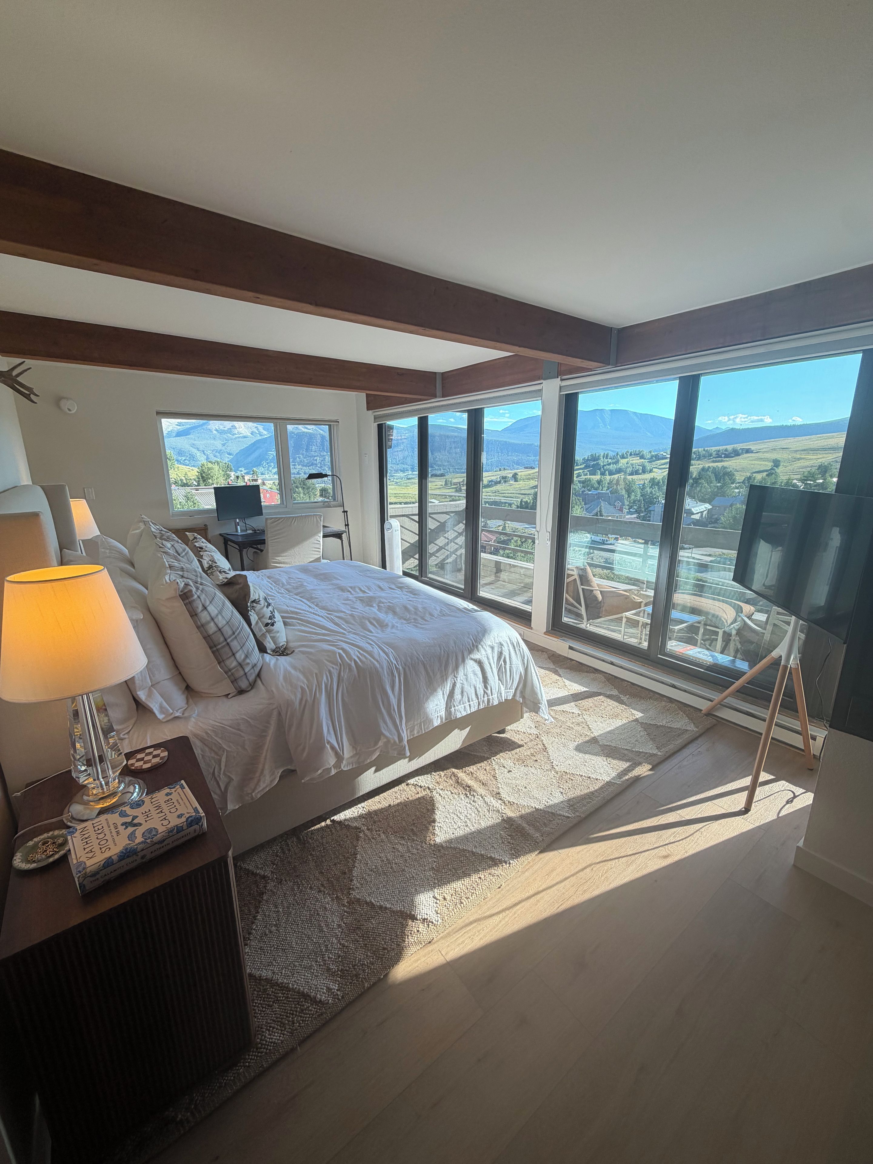 A bedroom with a large bed, a desk with a lamp, a chair, and a TV stand. There is a view of a mountainous landscape through large windows.