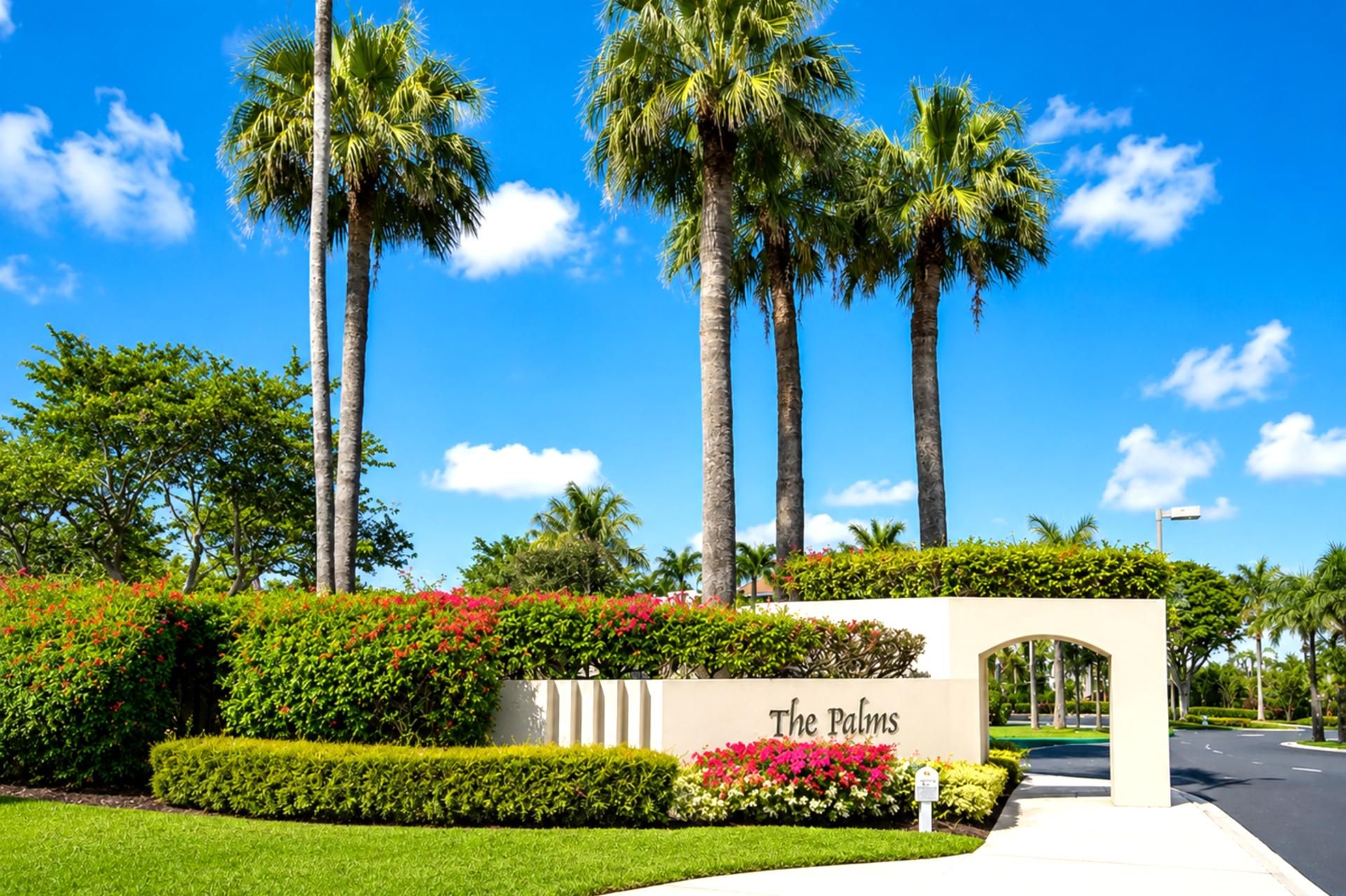 Une entrée aménagée avec des palmiers, une enseigne indiquant « The Palms », et une haie bien entretenue.