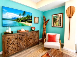 A living room with a wooden cabinet, a sofa, a coffee table, a large painting of a tropical beach, and a wooden sculpture. - NEW Palms at Wailea 2 Bedroom Garden-View Villa | Car Incl. w/ 6 nights | PAW-1706 by KBM (Lahaina)
