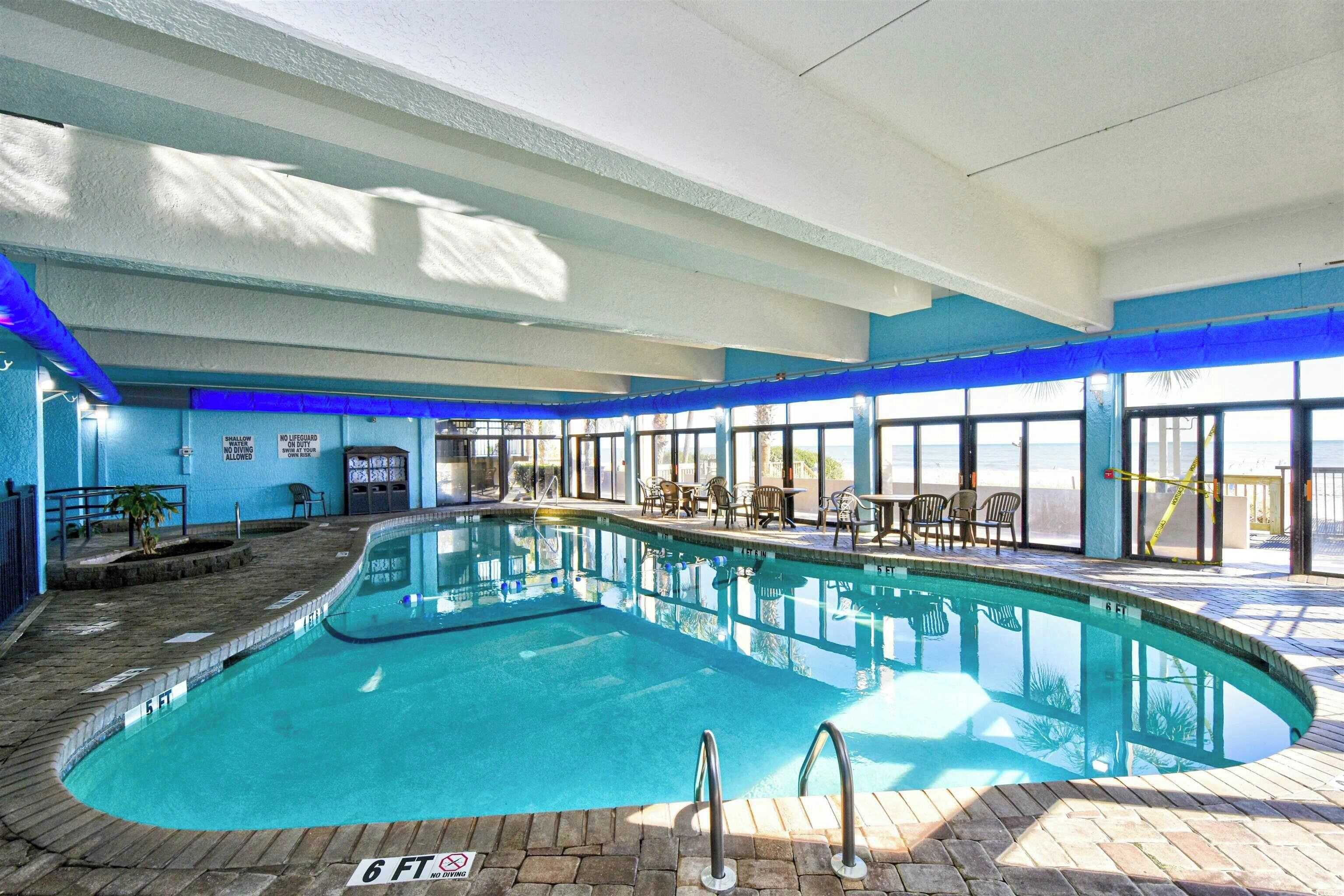 Una piscina interna con acqua cristallina, circondata da un solarium piastrellato e alcune sedie.