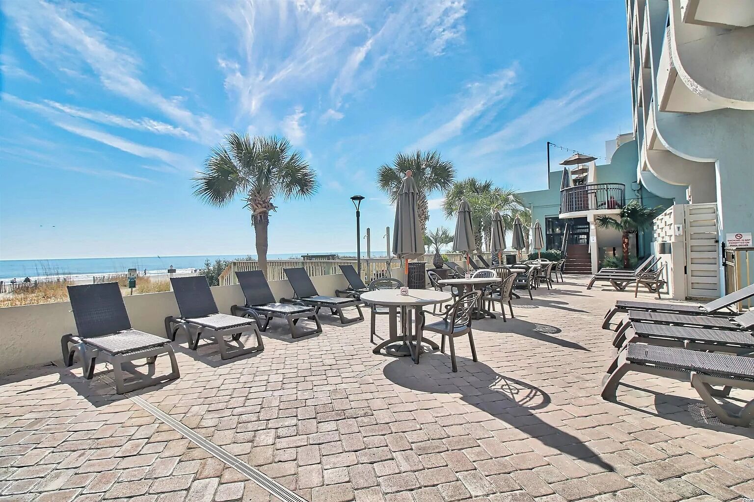 Ein sonniger Außenbereich mit Liegestühlen und Tischen mit Blick auf einen Strand und ein Gebäude mit Balkon.