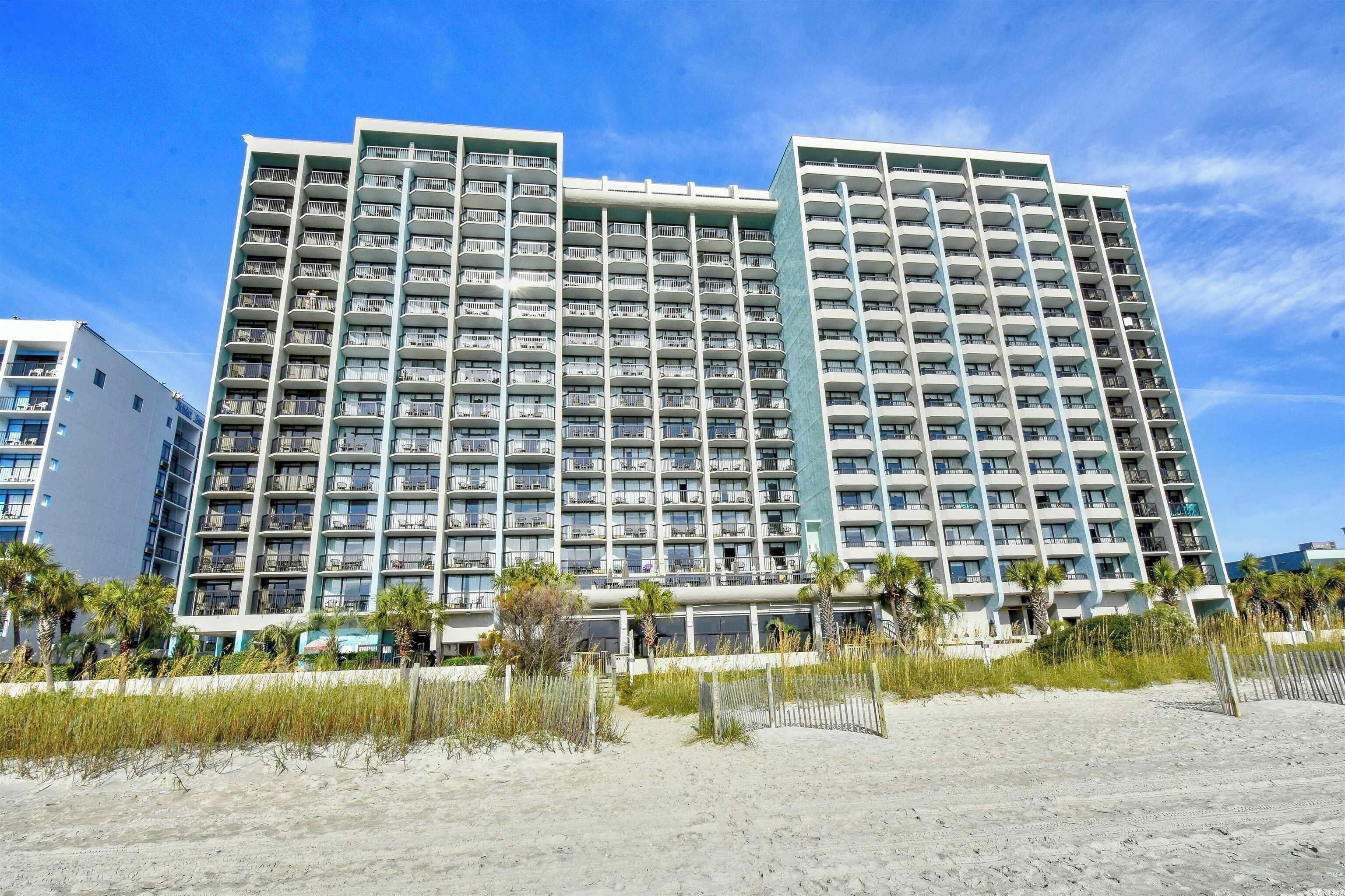 Un hôtel à plusieurs étages doté de nombreux balcons, situé près d’une plage de sable bordée de palmiers.