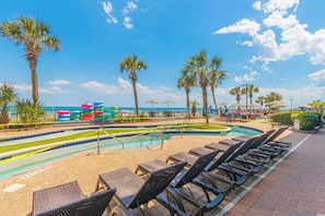 A pool area with lounge chairs, palm trees, and a water slide. - 2BR Oceanfront Full Kitchen free Waterprk sleeps 7 (Myrtle Beach)