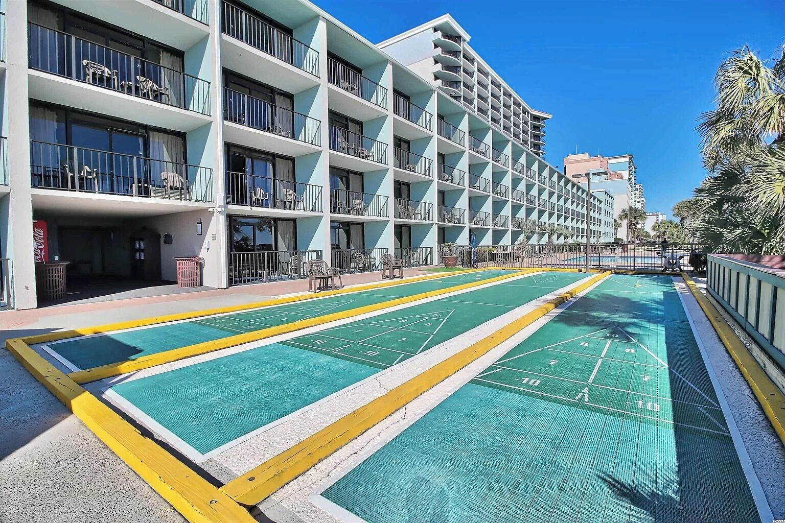 Ein Hotel mit mehreren Balkonen, einem Poolbereich mit grünen und gelben Markierungen sowie einem strahlend blauen Himmel.