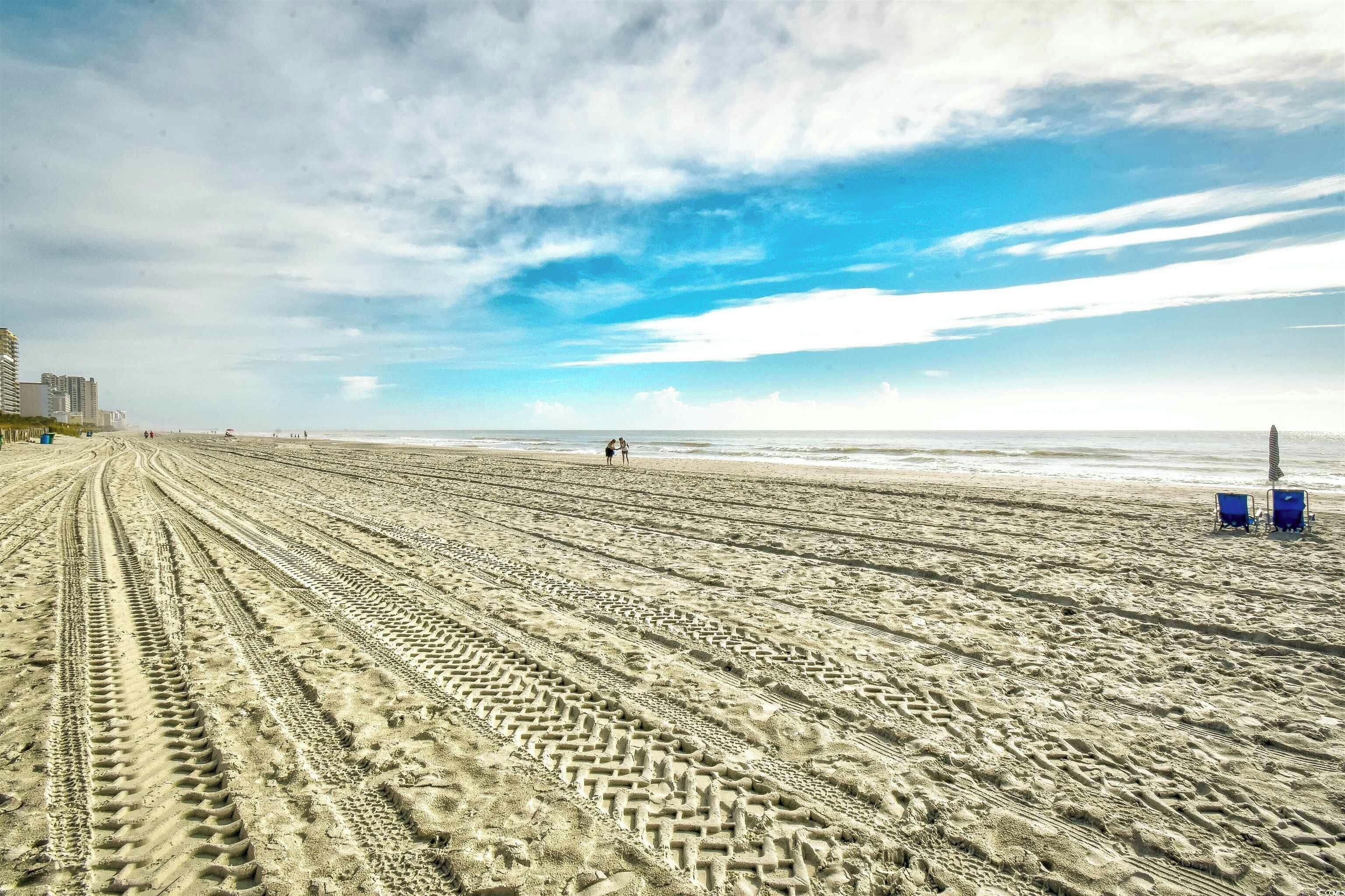 Ein weiter Sandstrand mit Radspuren, einigen Personen und einer entfernten Ansicht von Gebäuden.