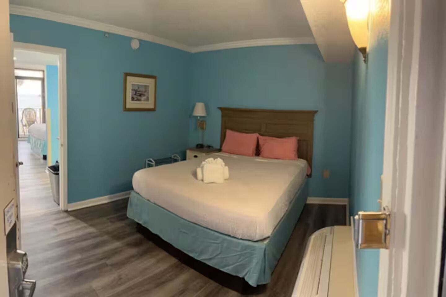 A bedroom with a bed, a nightstand, a lamp, a framed picture on the wall, and a door leading to another room.