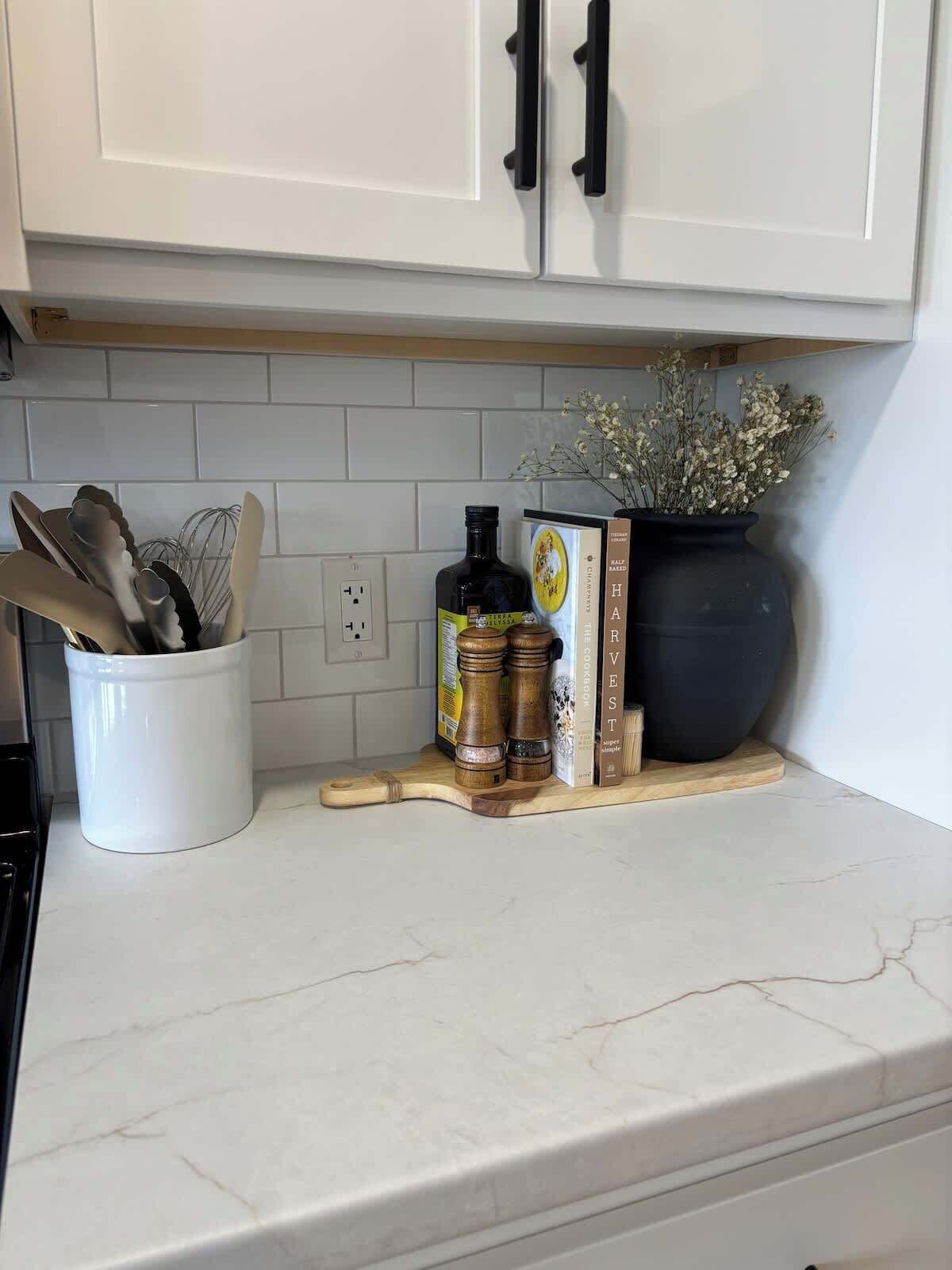 A kitchen with white cabinets, a white countertop, a white container with utensils, a wooden cutting board, a bottle of olive oil, a book, and a potted plant.