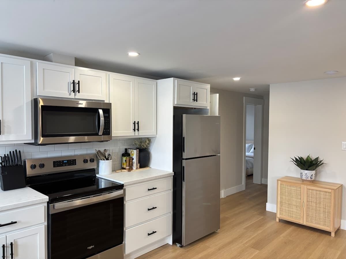 A modern kitchen with white cabinets, stainless steel appliances, and a wooden cabinet with a plant.