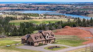 A large house with a driveway, a lawn, and a view of a lake and forest. - New Glasgow Pool House - Indoor Pool - Sleeps 14 (New Glasgow)