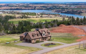 A large house with a driveway, a lawn, and a view of a lake and forest. - New Glasgow Pool House - Indoor Pool - Sleeps 14 (New Glasgow)