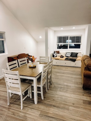 A dining area with a wooden table and chairs, a sofa with cushions, and a window with blinds.
